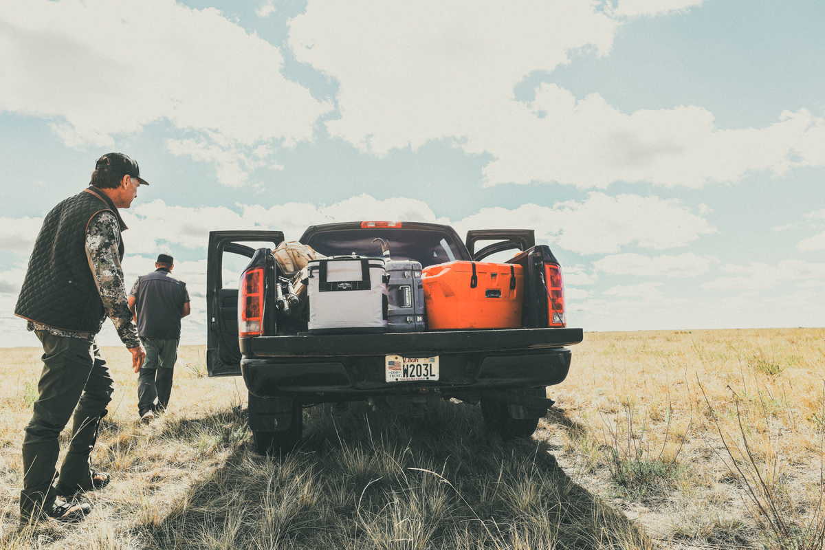 A black pickup truck parked in an open grassy field under a cloudy sky, with two men standing beside the open tailgate. Gear is loaded into the truck bed, including a orange cooler, a white bag, and other equipment.