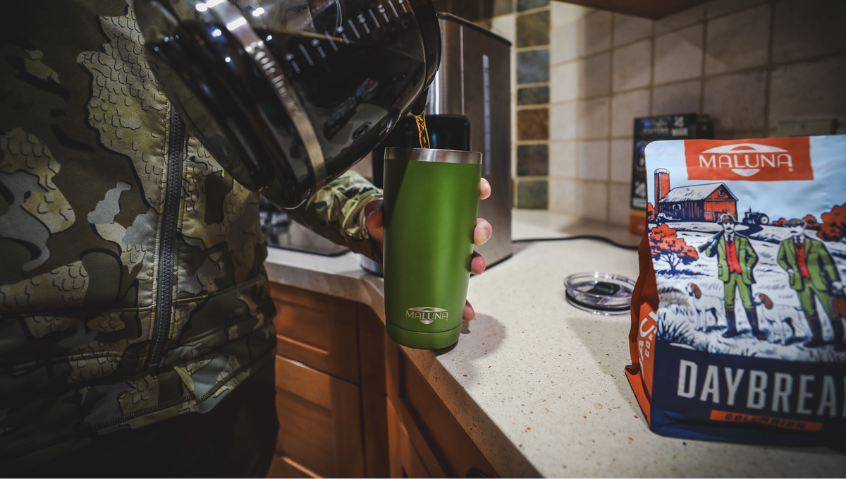 A person in a camouflage jacket pours hot water from a black coffee maker into a green insulated travel tumbler on a kitchen counter. A bag of Maluna Daybreak coffee is visible on the counter in the background.