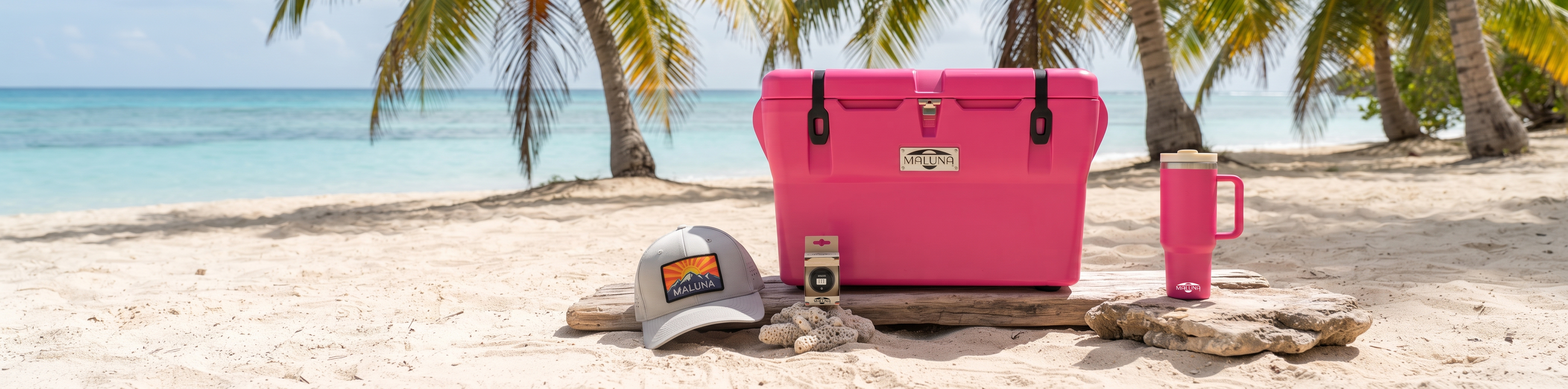 A bright pink cooler is set on a sandy beach with palm trees and the ocean in the background, alongside a matching pink handled mug, a gray cap, and a pink water bottle.