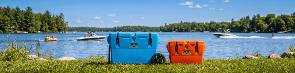 Two coolers, one blue with wheels and one orange, sit on grass by a lake. In the background, boats and people enjoy the sunny day on the water, with trees and blue sky.
