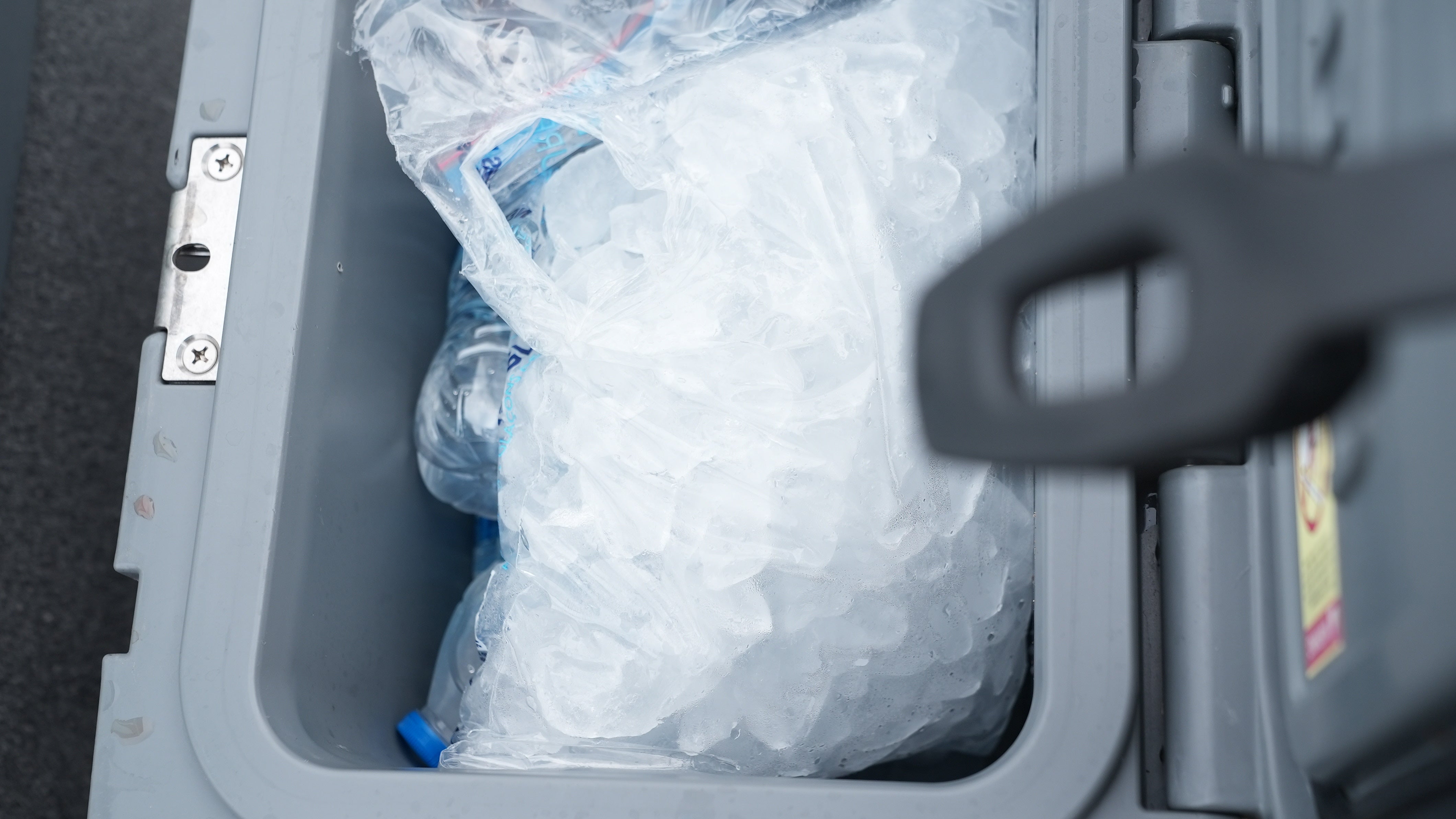 A close-up of a large plastic bag filled with ice cubes sits inside a gray cooler, with some water bottles partially visible beneath the ice.